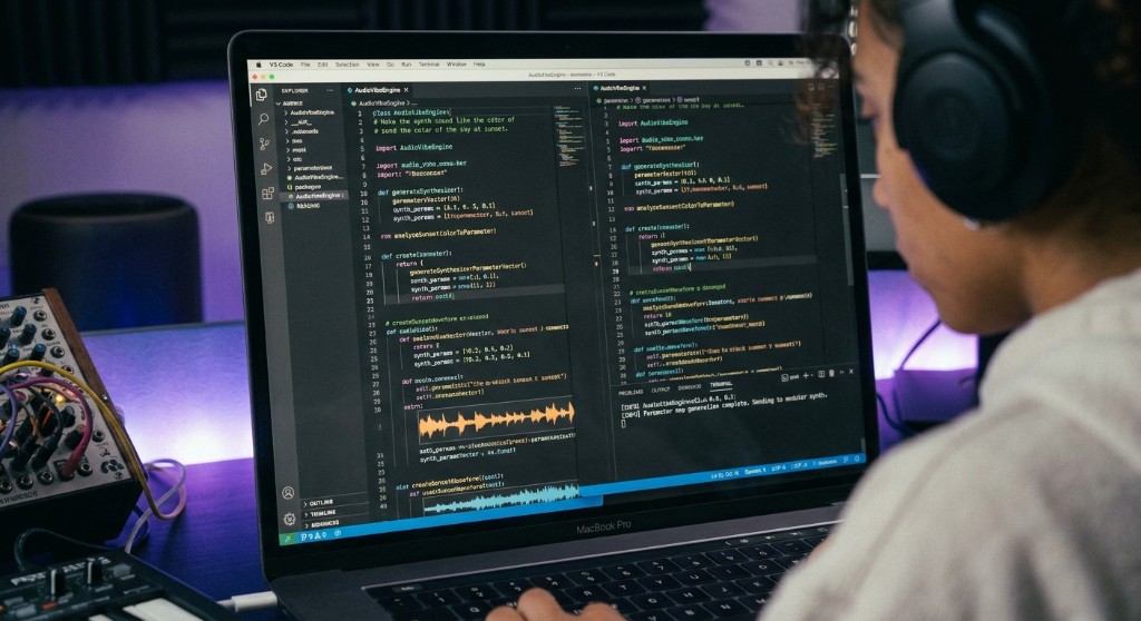 A person in headphones writes audio synthesis code on a laptop, with a synth and controller on the desk.